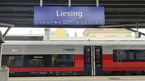 Im Bild zu sehen ein Foto vom Bahnhof Wien Liesing mit einem Zug im Hintergrund. Im Vordergrund sieht man auch das typische blaue Schild der Bahnhöfe mit der Beschriftung und entsprechenden Wegweisern zum Ausgang und zu Einrichtungen des Bahnhofes.
