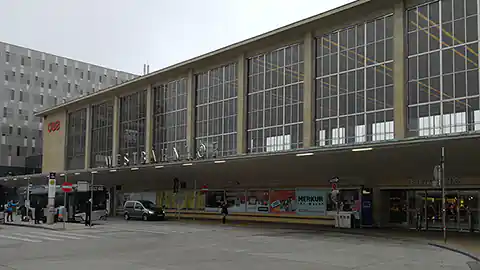 Die Vorderseite vom Bahnhof Wien Westbahnhof ist im Bild zu sehen. Diese Ansicht sieht man, wenn man vom Gürtel aus auf den Westbahnhof blickt, der Westbahnhof gehört zu den architektonischen Besonderheiten der Bahnhöfe in Österreich. Das Gebäude ist lang gezogen und hat eine riesige Glas-Fassade mit dazwischenliegenden Streben. Darunter sieht man ein Vordach des Bahnhofes, das den Eingangsbereich vor Regen schützt.