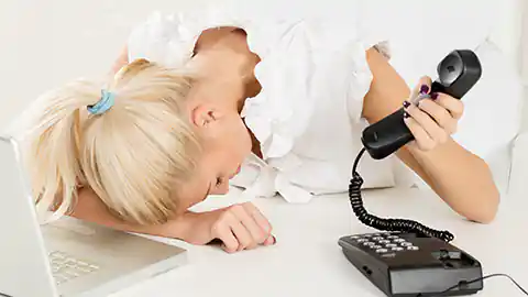 Eine blonde Frau, die in einem Büro arbeitet und dort eventuell Sekretärin oder Sachbearbeiterin ist, liegt mit dem Kopf auf ihrem Schreibtisch. Vor ihr ist ein Laptop aufgeklappt und in der Hand hält sie den Höhrer eines Festnetz-Telefones. Die Frau ist offensichtlich überfordert oder verärgert über die Arbeit. Sie ist jedenfalls erschöpft.