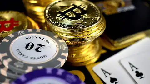 Spielkarten mit zwei Assen sowie mehrere Chips aus einem Casino liegen neben einem Stapel von Bitcoin-Münzen. Die Münzen sind Kryptowährung.
