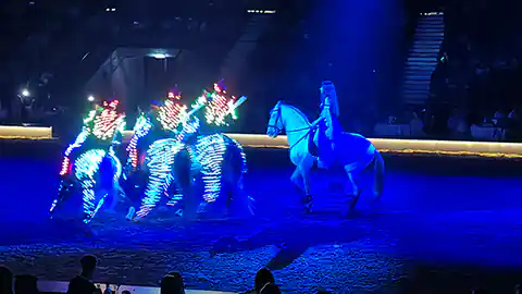 Szene aus der Cavalluna Pferdeshow mit drei Reitern die in bunten Leuchtkostümen mit Led-Lampen am ganze Körper nebeneinander reiten. Daneben steht im blauen Licht angestrahlt eine weitere Reiterin mit einem weißen Pferd.