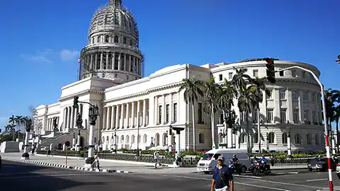 Unter einem strahlend blauen Himmel ist das Parlamentsgebäude von Kuba in Havanna zu sehen. Es ist ein lang gezogenes Haus mit vielen Säulen und in der Mitte mit einem hohen Turm und einer runden Kuppel am Dach.