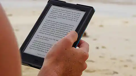 An einem Sandstrand benutzt eine Person einen E-Book-Reader zum Lesen. Man sieht im Bild den Reader mit Text darauf und die rechte Hand der Person, die ihn hält. Sonst ist nur der Sandstrand im Hintergrund zu sehen. Der Blick geht am rechten Oberarm der Person vorbei, die den E-Book-Reader zum Lesen des Buches benutzt.