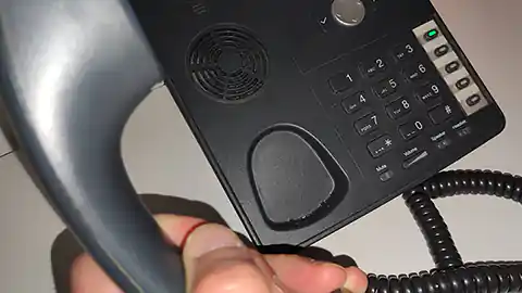 Ein Festnetztelefon mit Tasten, das abgehoben wurde. Der Apparat ist etwas moderner, hat ein paar Zusatzfunktionen und ist schwarz. Der Telefonhörer wird von einer Hand gehalten, die teilweise im Bild zu sehen ist.
