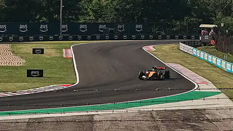 Ein Rennauto auf einer Formel-1-Rennstrecke fährt gerade in eine Kurve ein. Im Bild ist das Rennauto zu sehen und im Hintergrund der Verlauf der Rennstrecke sowie eine Seitenbande mit Werbung darauf und am Horizont ist Wald zu erkennen.