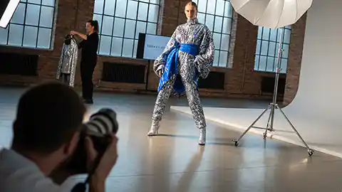 In einem Fotostudio steht ein Model neben einem Reflektor. Das Model ist in einem ungewöhnlichen Overwall mit einer blauen Schleife an der Taille gekleidet. Im Vordergrund des Bildes kniet ein Fotograf auf dem Boden um ein Bild von dem Model zu machen. Das Bild zeigt eine Perspektive, bei der man mit der Kamera über die Schulter des Fotografen auf das Model blickt. Neben dem Model geht eine Stylistin mit einem Kleid im Hintergrund noch aus dem Blickfeld der Kamera.