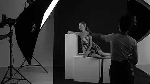 Schwarz-Weiß-Aufnahme in einem Fotostudio. Umgeben von Blitzlicht-Schirmen und Beleuchtung ist ein Fotomodel auf einigen zusammengestellten weißen Kisten zu sehen. Davor steht eine andere Person, die gerade die Szene erklärt.