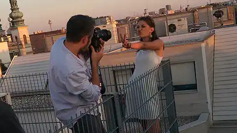 Auf einer Dachterrasse in Wien findet ein Fotoshooting statt. Ein Fotograf im Vordergrund macht Aufnahmen von einem Model, das sich an das Geländer der Dachterrasse lehnt und ihm die linke Hand mit einem Viktory-Zeichen entgegen streckt. Im Hintergrund sind weitere Häuserdächer zu sehen und ein Kirchturm. Das Bild wurde auf einer Dachterrasse im 1. Bezirk in Wien gemacht.