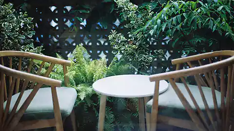 In einem Garten stehen zwei Stühle und ein kleiner Tisch auf einer Terrasse. Der runde Tisch ist niedrig, die beiden Stühle sind aus Holz. Dahinter sind Pflanzen zu sehen.