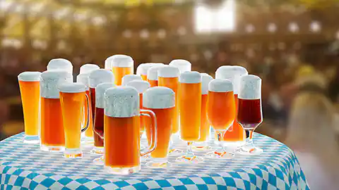 Auf einem Tisch mit einer blau-weiß karierten Tischdecke stehen viele Bierkrüge die mit Bier gefüllt sind. Das Bild ist auf dem Oktoberfest entstanden und symbolsiert ein Volksfest, im Hintergrund ist eine Halle zu erkennen, in der das Publikum ist.