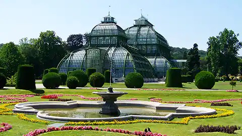 Das Palmenhaus in Wien ist ein Gebäude aus viel Glas. Es ist mit mehreren Kuppeln ausgestattet und steht in einem Park, der im französischen Stil gehalten ist. Die Blumen und Sträucher sind sehr streng nach geometrischen Formen zugeschnitten. Der Himmel ist hellblau.