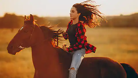 A female rider is galloping bareback on her brown horse through a summery landscape. The horse is wearing a bridle, but the rider is riding without a saddle. She is wearing ripped blue jeans and a red-and-black plaid top. She has long brown hair that is blowing in the wind. The landscape in the background has a steppe-like appearance. Only the horse's head, neck, and upper body are visible in the image.