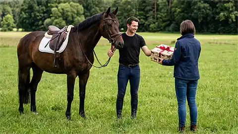 Ein Mann in dunkler Kleidung steht neben seinem Pferd auf einer Wiese. Mit der rechten Hand hält er das aufgesattelte Pferd am Zügel, mit der linken Hand nimmt er das Geschenkpaket einer anderen Person entegegen, die im Bild mit dem Rücken zur Kamera steht. Im Hintergrund ist nur die große Wiese und ein Wald zu sehen.