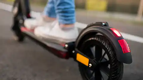 Detailaufnahme eines Scooters, eines sogenannten Elektro-Rollers mit zwei kleinen Rädern. Auf dem Scooter steht eine Person, die weiße Sneakers und blaue Jeans trägt. Der Scooter hat ein Rücklicht.