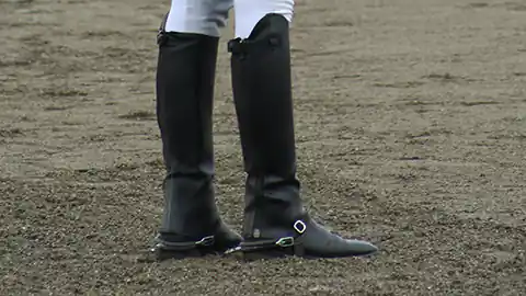 Ein Reiter steht auf einem Sandplatz. Man sieht in der Detailaufnahem die Reitstiefel mit Sporen. Es sind schwarze Reitstiefel und die Person trägt eine weiße Reithose mit hellgrauem Lederbesatz.