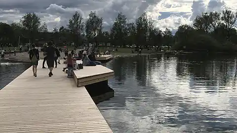 Im Bild zu sehen ist ein Holzsteg, der sich an einem Badeplatz / Strandbad an einem See befindet. Auf dem Steg sind einige Menschen zu sehen, das Bild wurde vom Steg in Richtung Seeufer aufgenommen. (Das konkrete Bild zeigt das Strandbad in Seekirchen am Wallersee.)