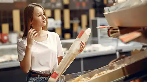 Eine junge Frau mit langen braunen Haaren und einem weißen T-Shirt steht in einem Supermarkt in der Backabteilung. Sie hält ein Baguette in der Hand und blickt auf die vor ihr liegenden Regale mit Brot und Gebäck.