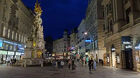 Eine Abendaufnahme aus dem Zentrum der Stadt Wien. Zu sehen ist die Pestsäule am Wiener Graben umrahmt von zahlreichen Geschäften und Lokalen und vielen Passanten, die sich am Abend hier bewegen.