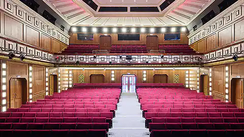 Innenraum des Theater Akzent mit der markanten Architektur und der markanten Wandverzierung ist in diesem Bild zu sehen. Das Theater in Wien hat links und rechts zwei Blöcke mit samtroten Stühlen für das Publikum. In der Mitte ist ein langer Gang nach vorne und in der Bildmitte sieht man dann die Bühne.